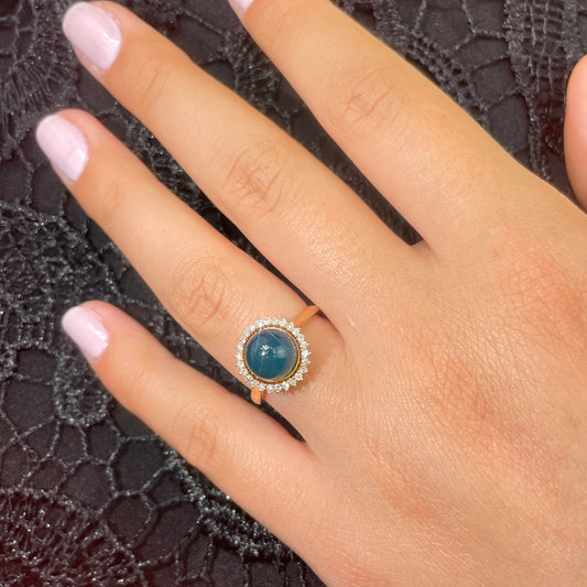 Hand wearing a ring with a blue amber gemstone and diamonds on a black background