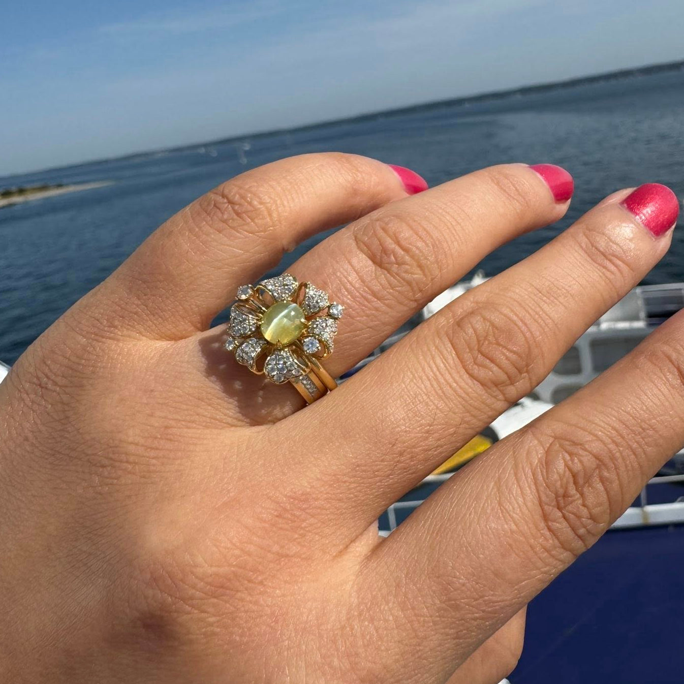 Hand wearing a decorative ring with a cat's eye gemstone against a blue sky and water background.
