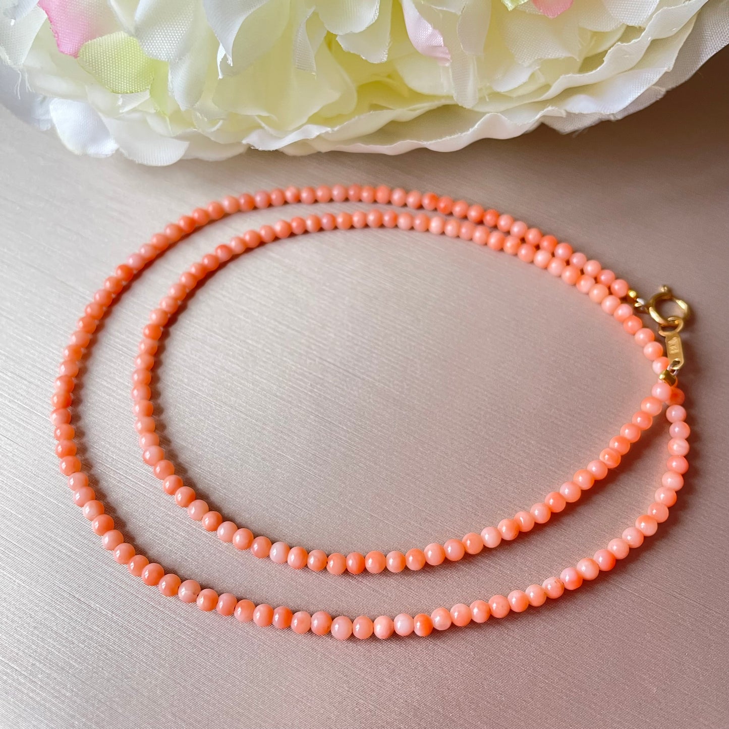 Coral beaded necklace with a gold clasp on a light pink surface with flowers in the background.