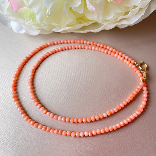 Coral beaded necklace with a gold clasp on a light pink surface with flowers in the background.