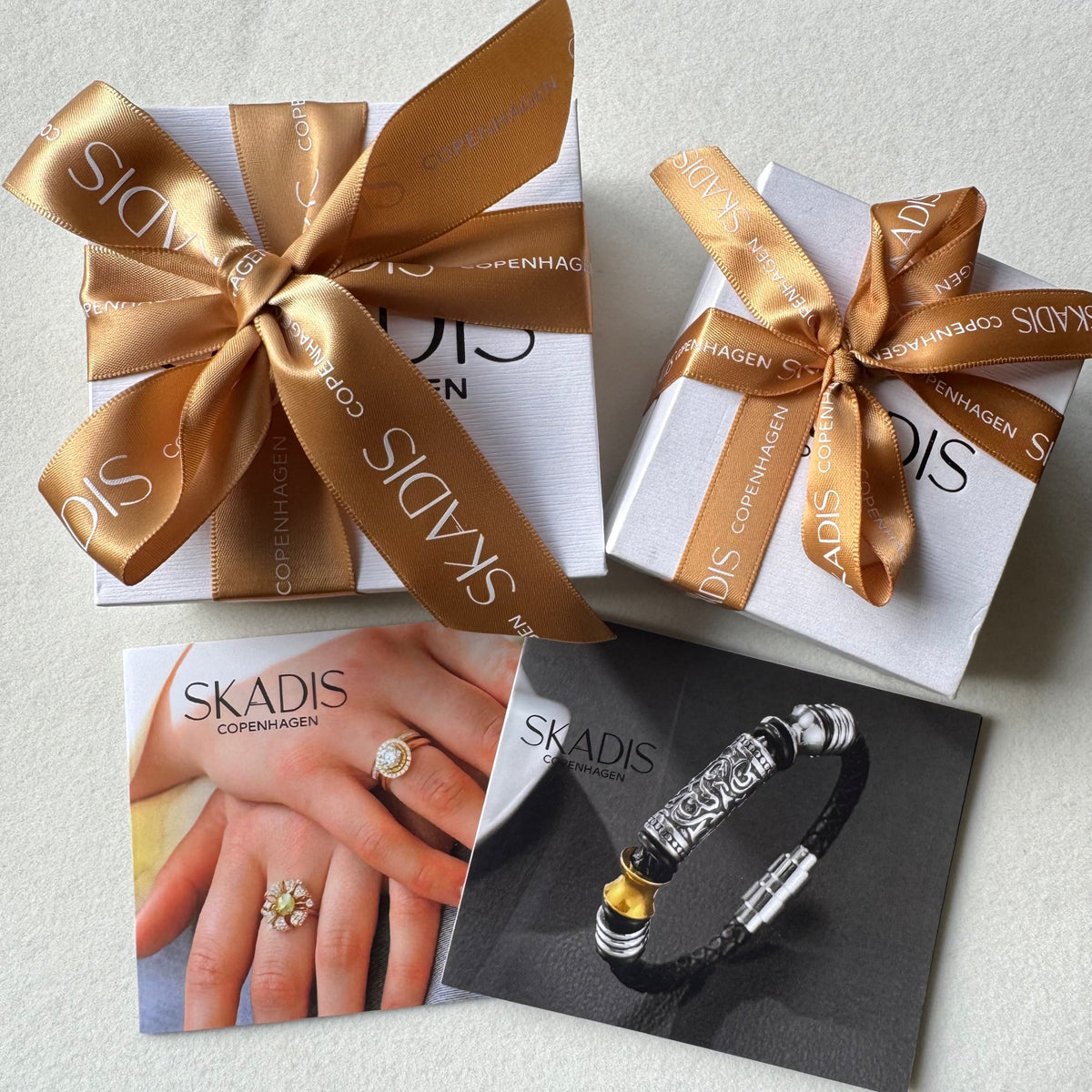 Three SKADIS gift boxes tied with metallic gold ribbons are arranged on a light surface, alongside printed cards showing a ring on hands and a bracelet.