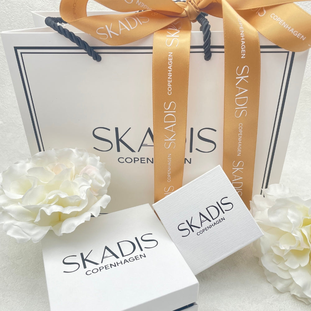 SKADIS Copenhagen gift packaging set featuring a white shopping bag with a gold ribbon, a white gift box, and a smaller branded card, styled with white flowers on a light background.