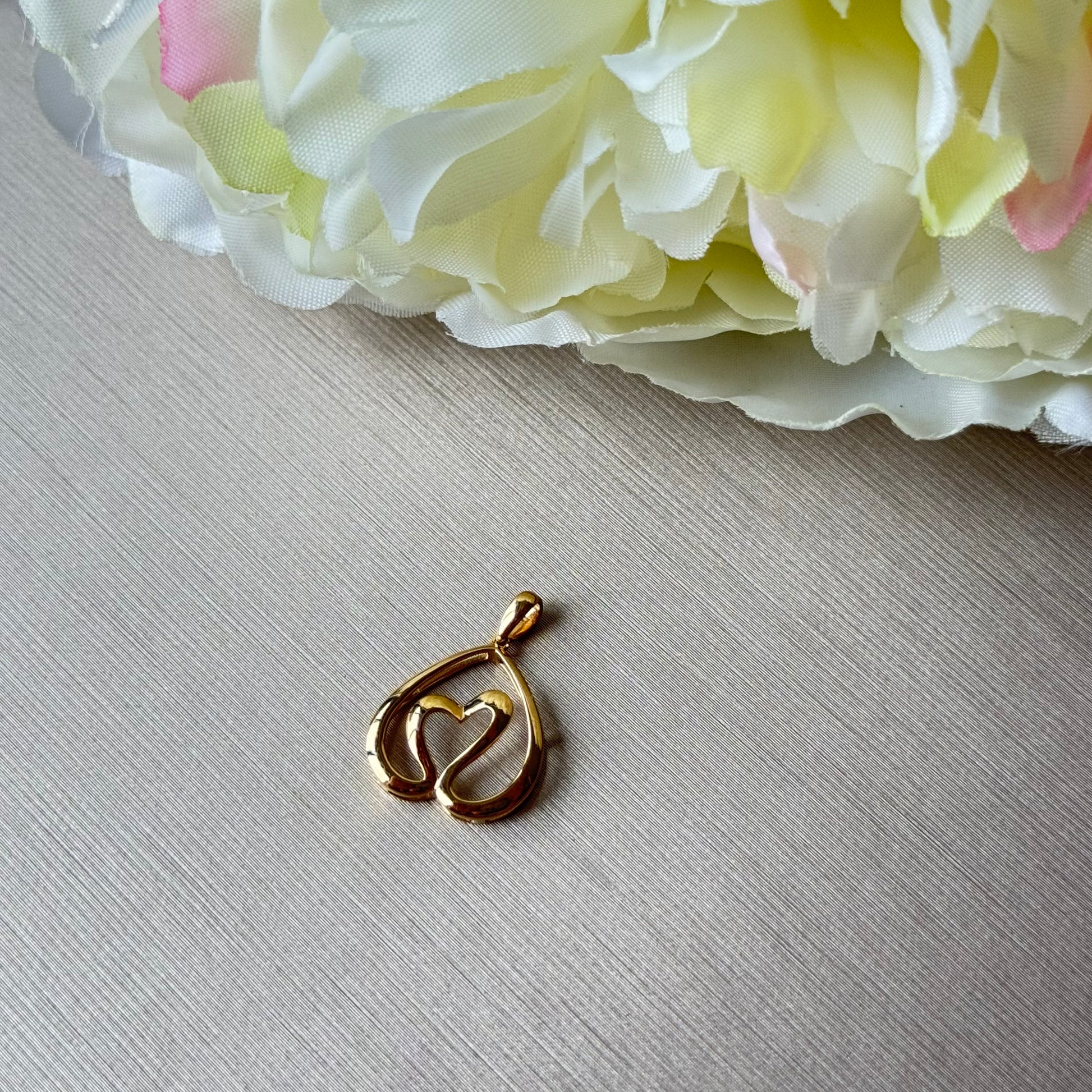 Gold pendant with heart design on a textured surface with flowers in the background.