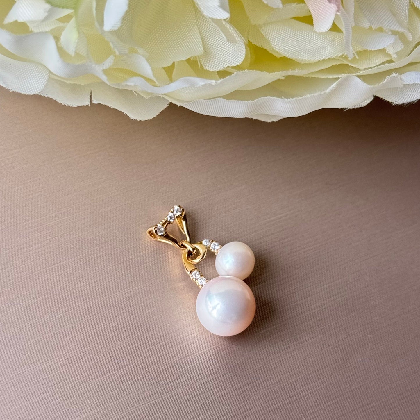 Pearl pendant with gold setting on a beige surface with flowers in the background.