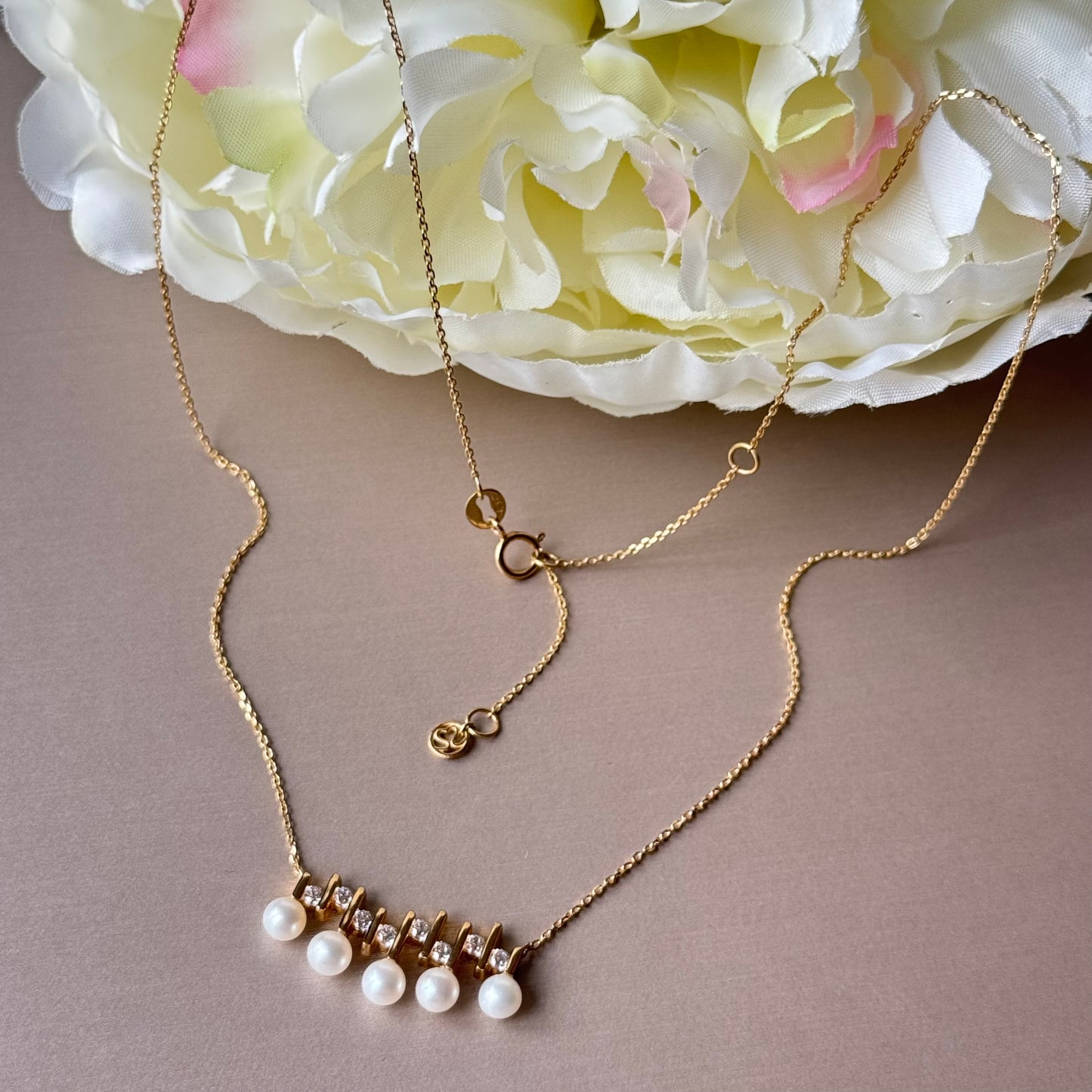 Gold necklace with pearl drops on a beige surface with flowers in the background.