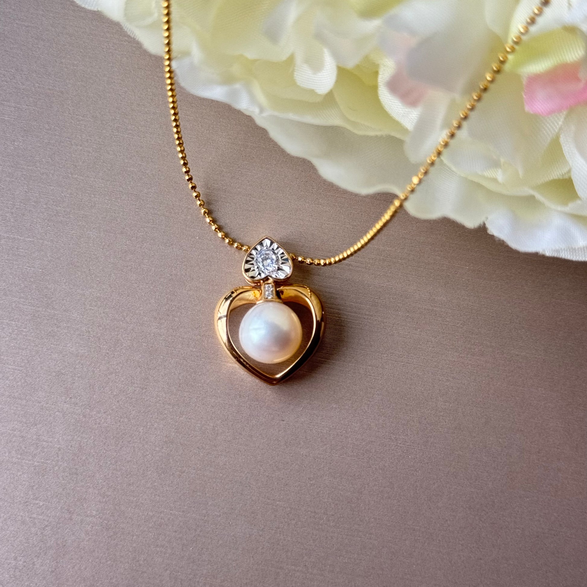 Gold necklace with a heart-shaped pendant featuring a pearl against a soft pink background.